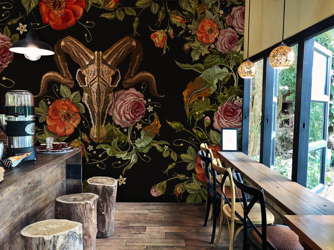 Dark floral skull wallpaper with roses, rustic wooden flooring, modern cafe setting, large windows, stools.