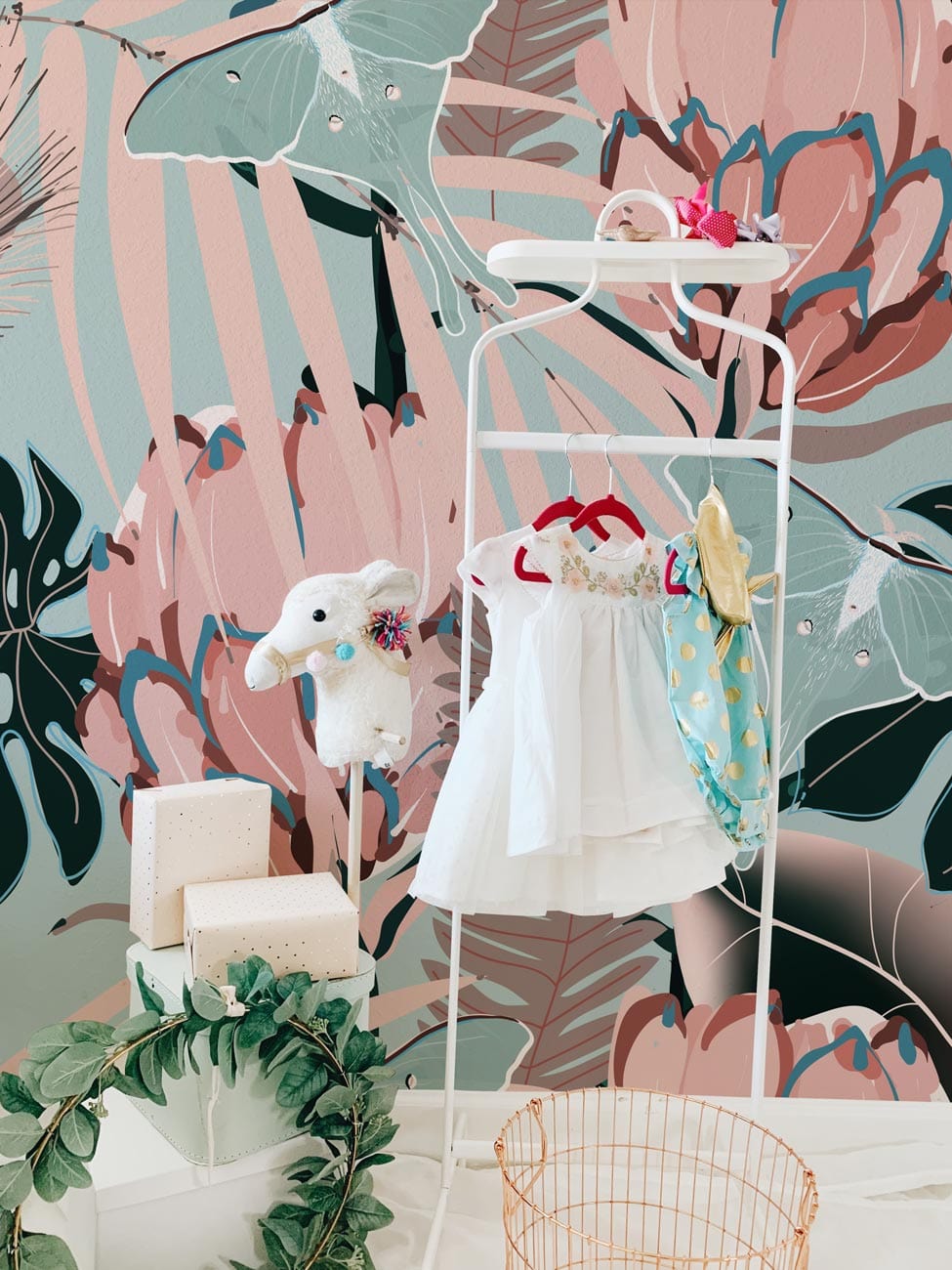 Alt text: Vintage tropical floral wallpaper in nursery with white clothing rack, soft toys, and pink decor.