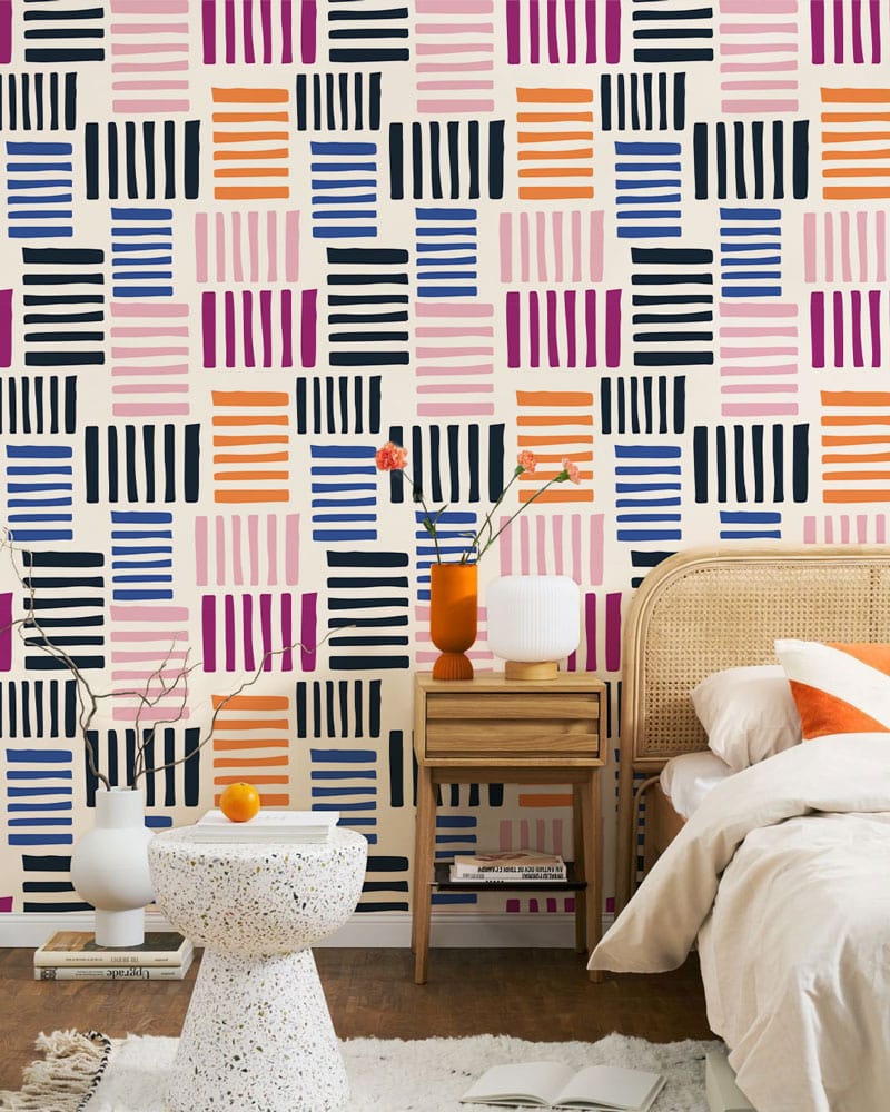 Vibrant geometric stripe wallpaper in pink, orange, blue. Bedroom with rattan bed and wooden nightstand.