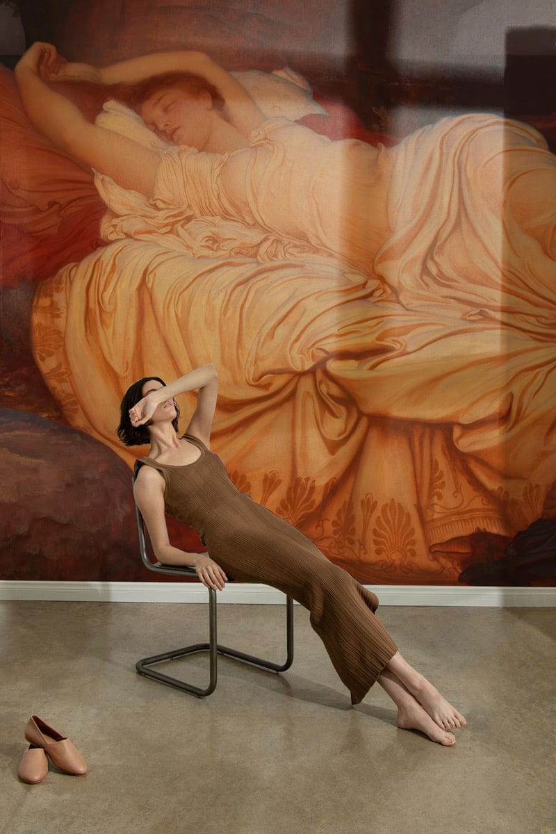 Woman lounging before Elegant Victorian mural wallpaper in earthy tones, minimalist chair, concrete flooring.
