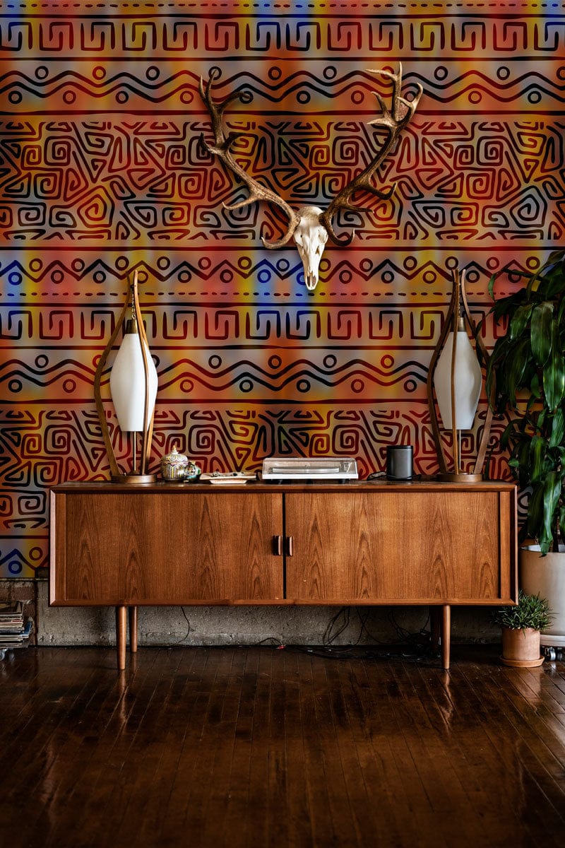 Vibrant tribal wallpaper with orange-blue hues, mid-century wooden sideboard, mounted stag horns, hardwood flooring.