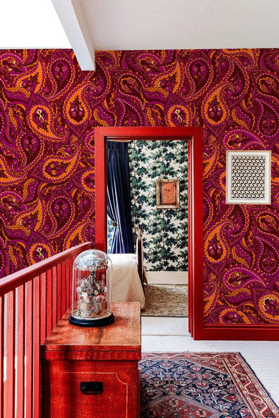 Vibrant paisley wallpaper in burgundy tones, red stair railing, hallway, wooden furniture, patterned rug.