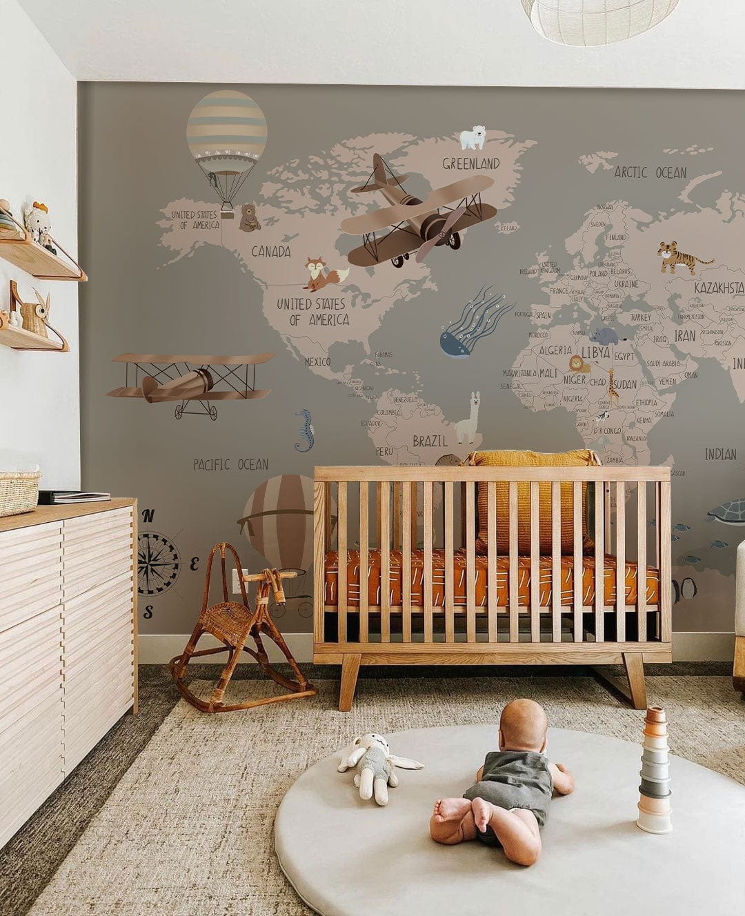 Vintage World Map Mural Wallpaper in nursery with aeroplanes, crib, taupe tones, wooden furniture, rug.
