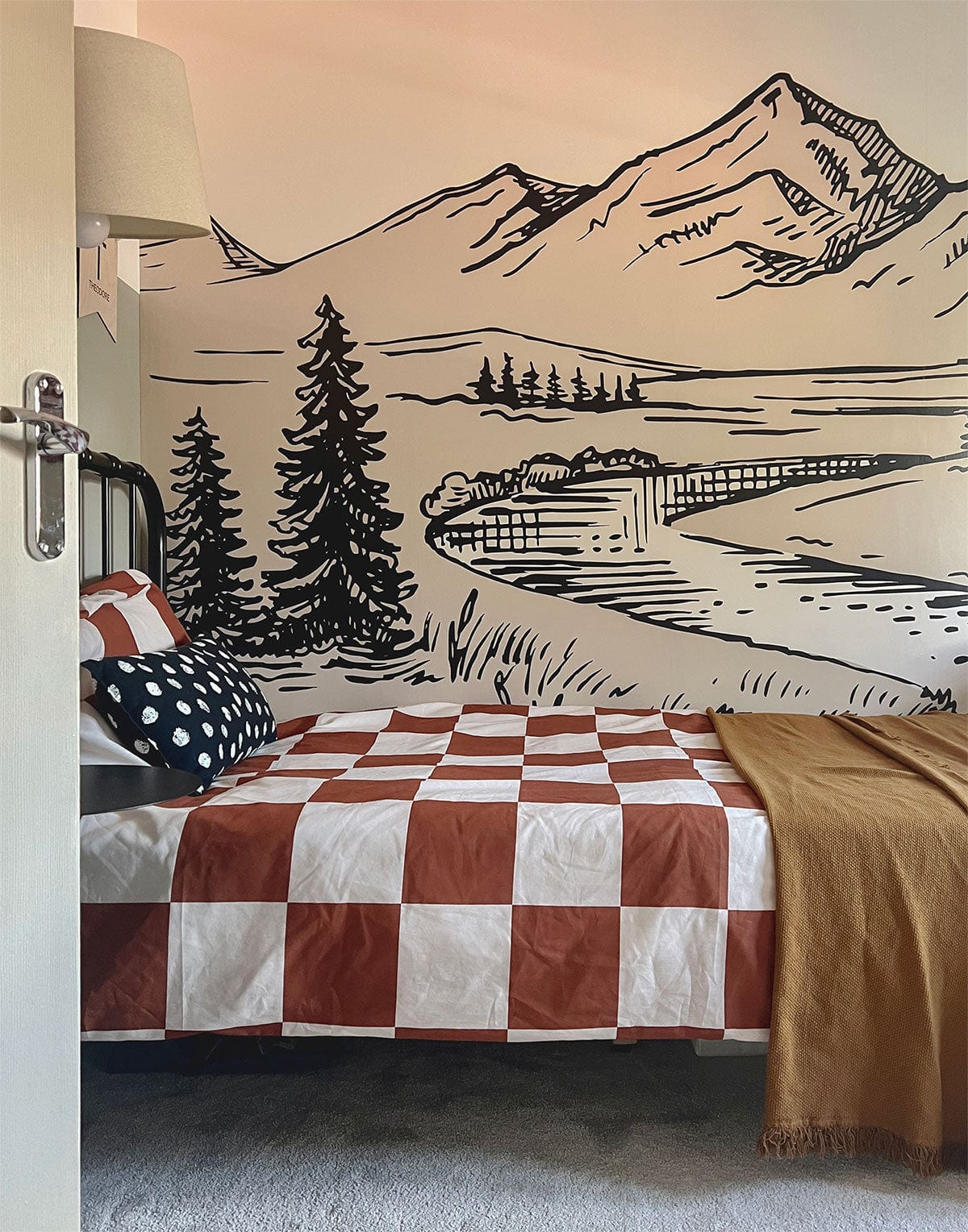 Beige and black landscape mural wallpaper with bed, checkered duvet, and bedside lamp in bedroom.