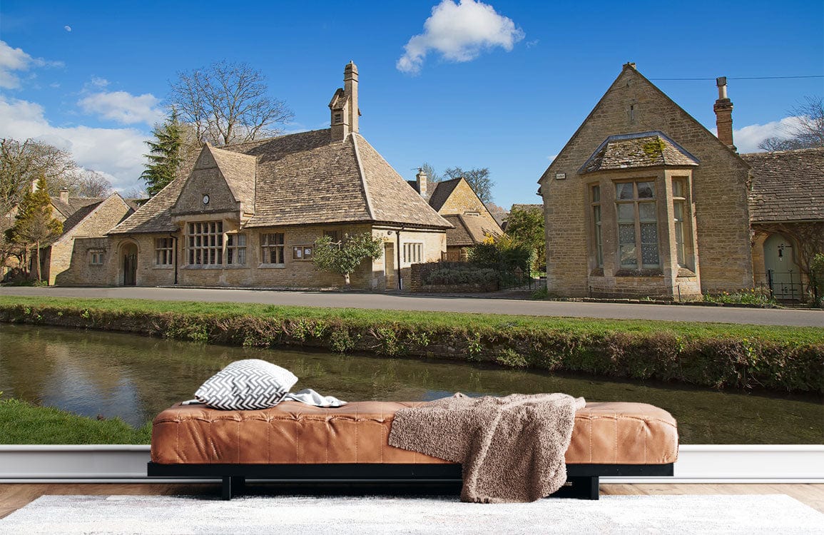 Charming Cotswold Village Scene Mural Wallpaper, warm stone hues, brown leather sofa, light wooden flooring.
