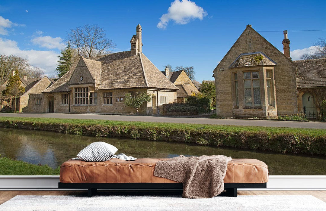 Charming Cotswold Village Scene Mural Wallpaper, warm stone hues, brown leather sofa, light wooden flooring.