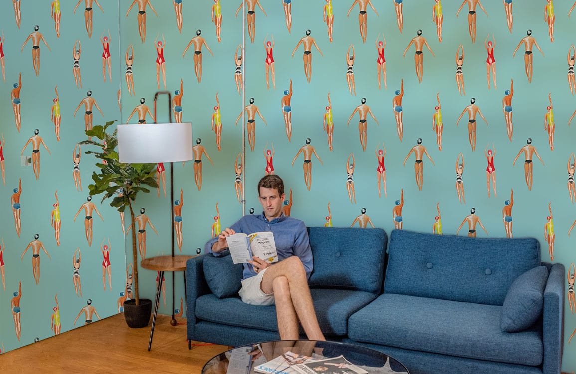 Man reading on retro blue sofa, aqua wallpaper; wooden floor; Retro Swimmers patterned beach decor.