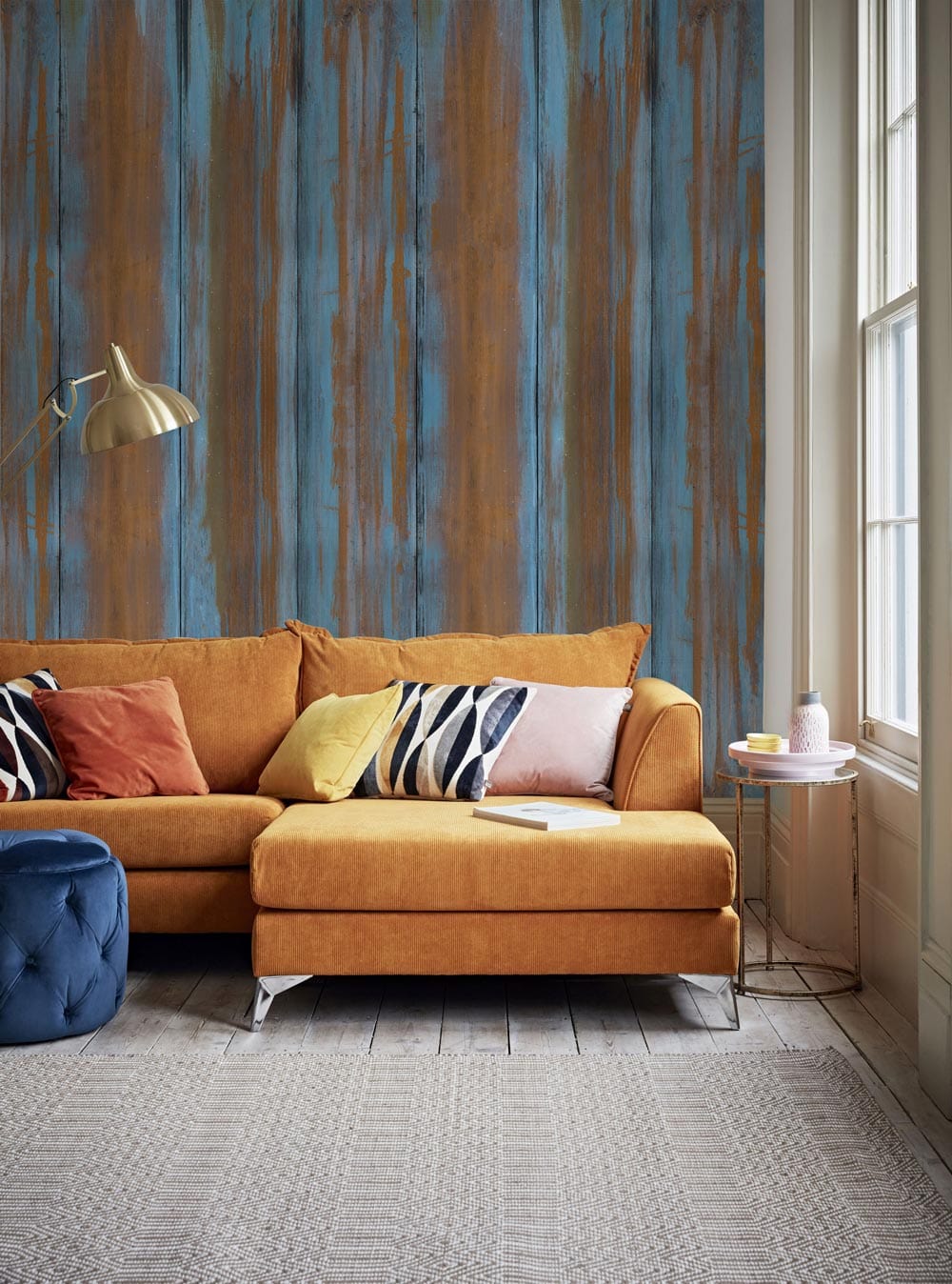 Rustic copper and blue striped wallpaper in living room with mustard sofa, blue ottoman, wood flooring.