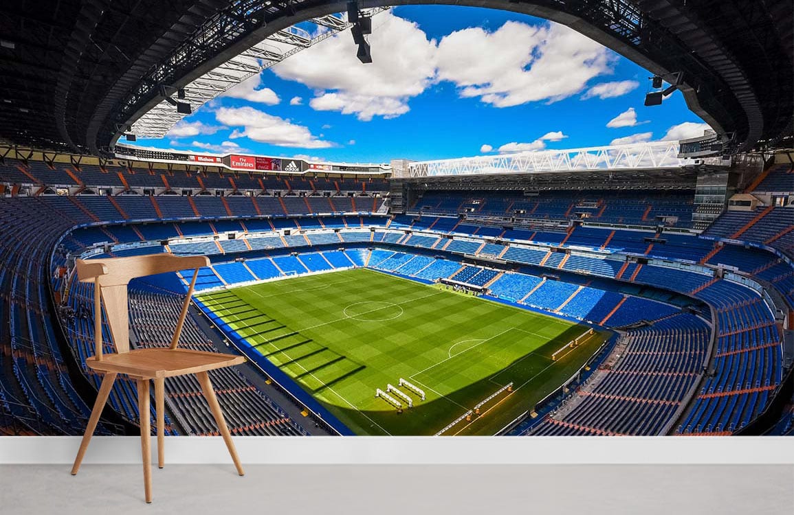 Vibrant football stadium mural wallpaper with green pitch, blue seats, wooden chair, grey flooring.