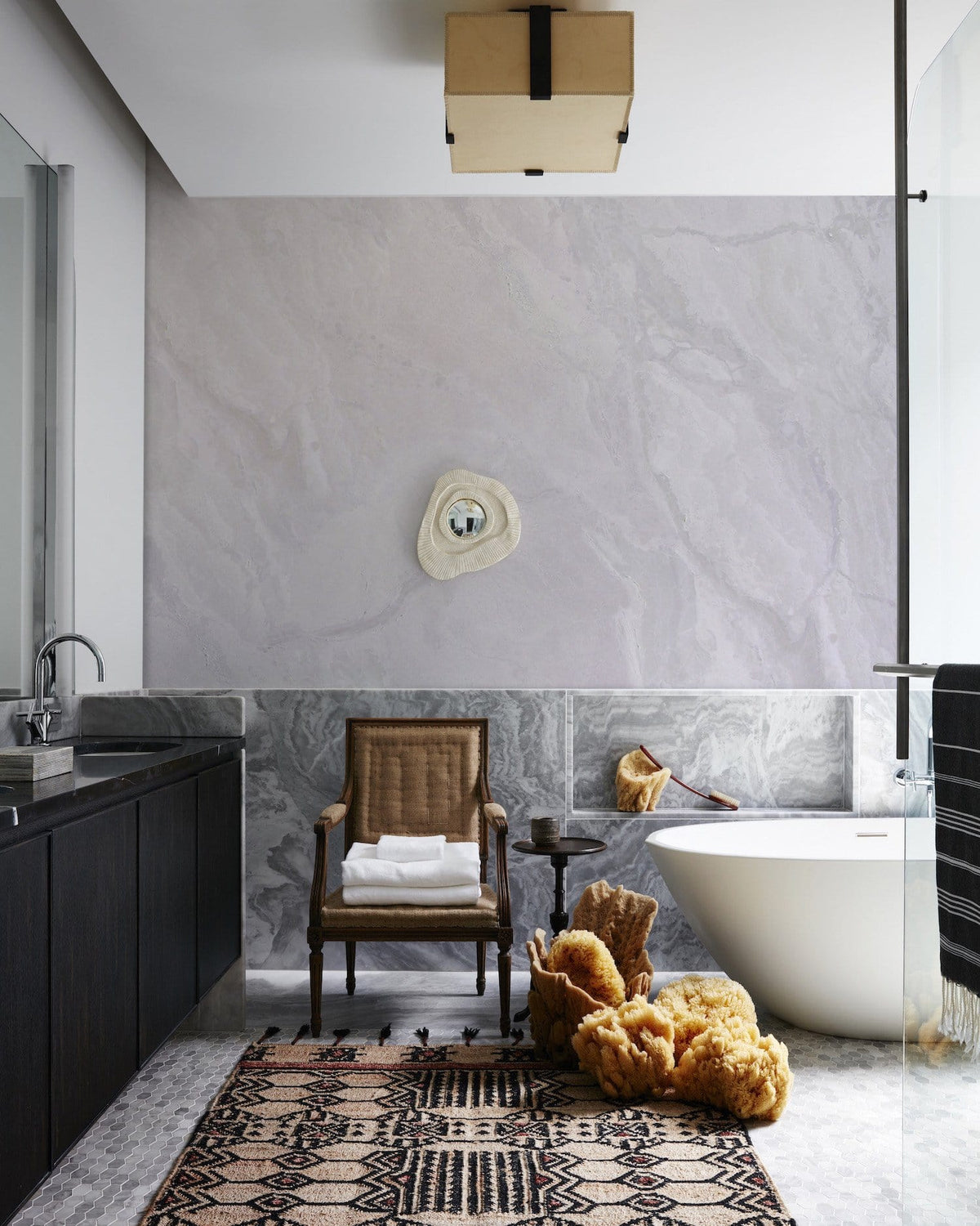 Bathroom with elegant marble effect mural wallpaper, dark vanity, freestanding bathtub, and woven chair.
