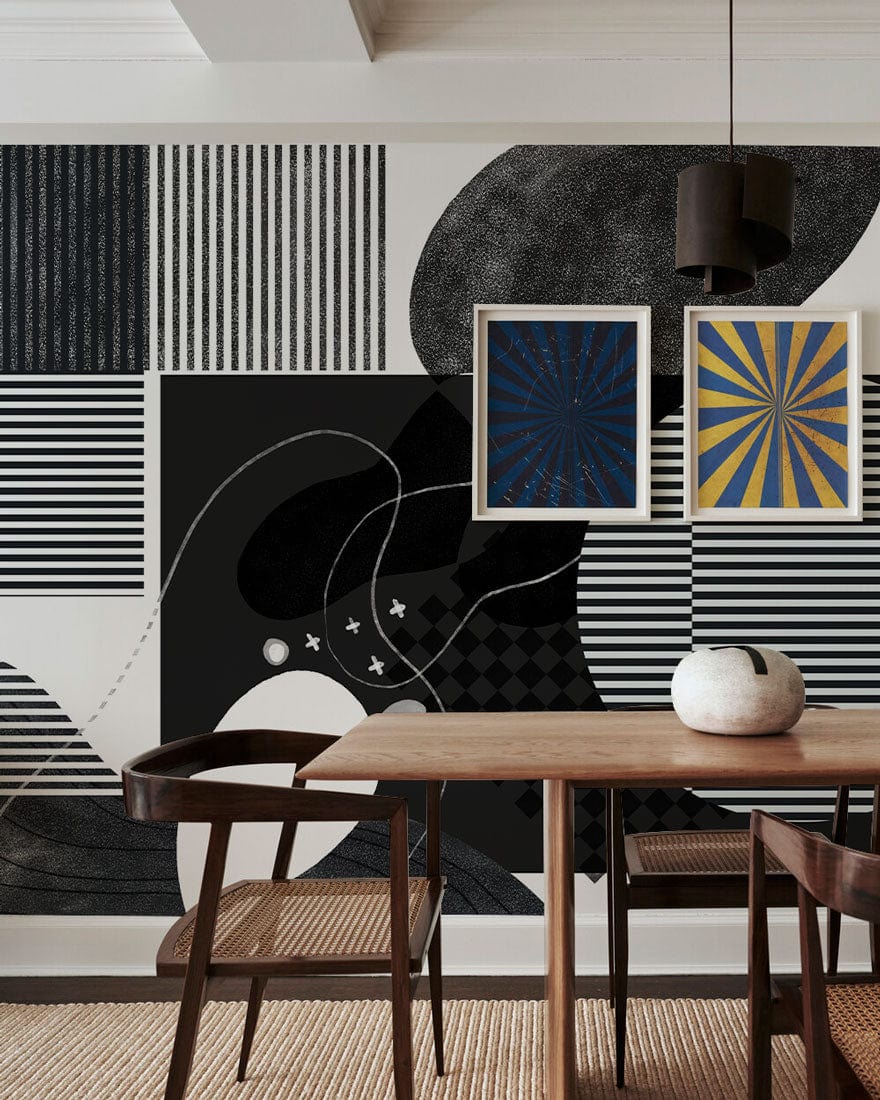 Monochrome abstract geometric wallpaper in dining room with wooden table, black pendant light, and art prints.