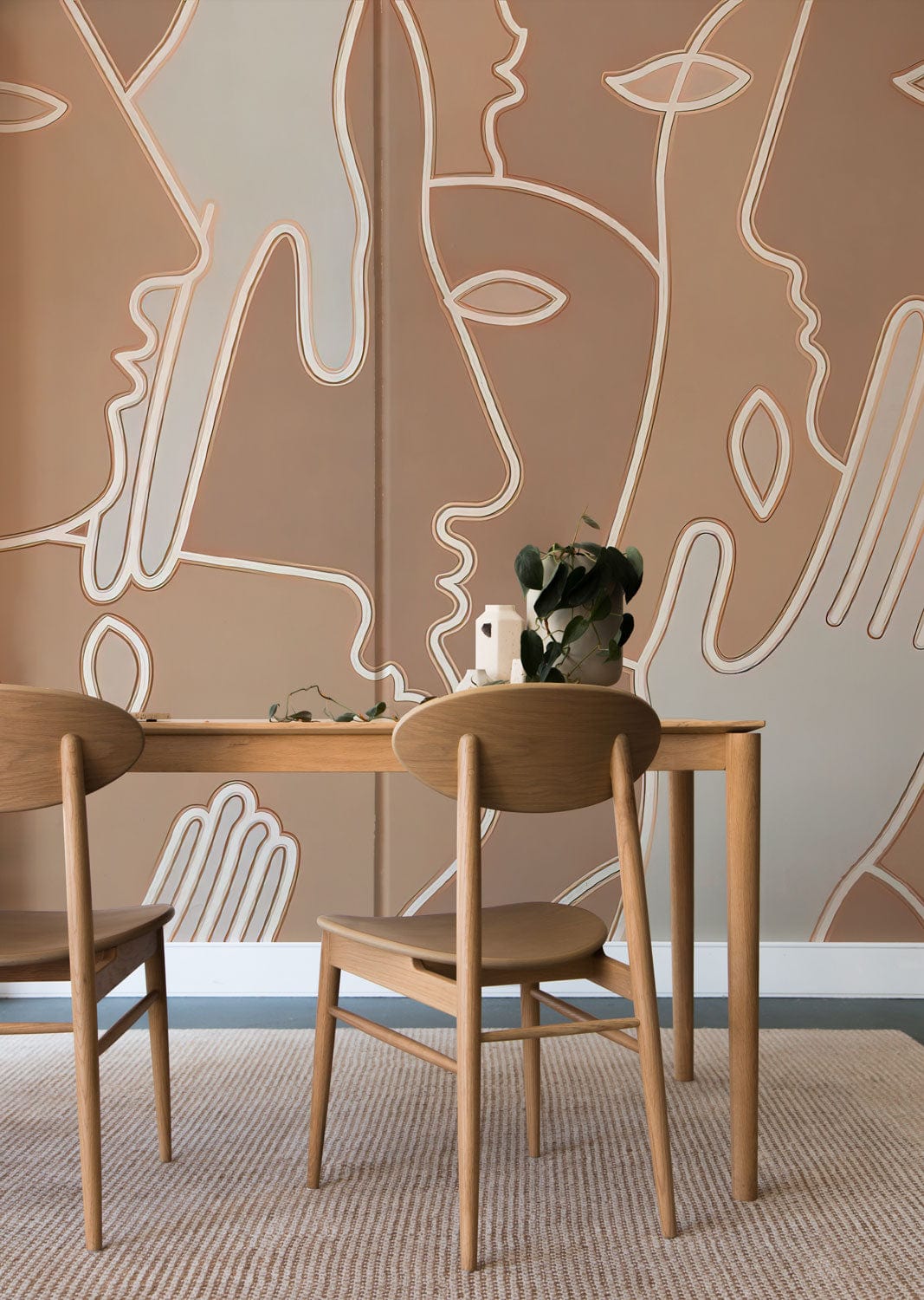 Beige line art wallpaper with abstract faces, wooden dining table, chairs, and light carpet flooring.