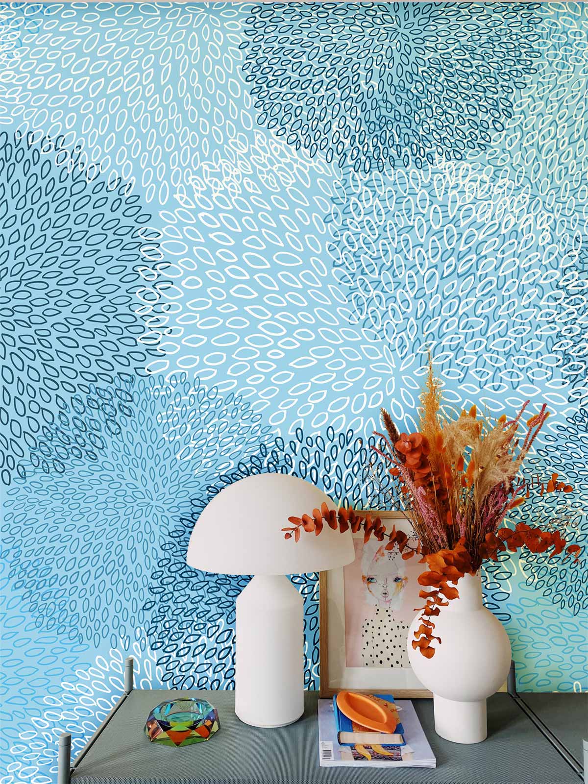 Elegant blue floral wallpaper with modern oceanic design; white lamp, decorative vases on table.