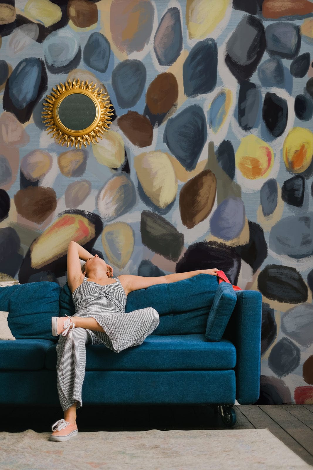 Elegant room with multicoloured pebble mural wallpaper, teal sofa, gold mirror, dark wood flooring.