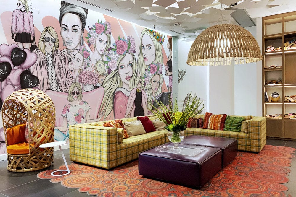 Chic Fashionista mural with pastel pink illustrations, plaid sofas, modern pendant lighting, vibrant patterned rug.