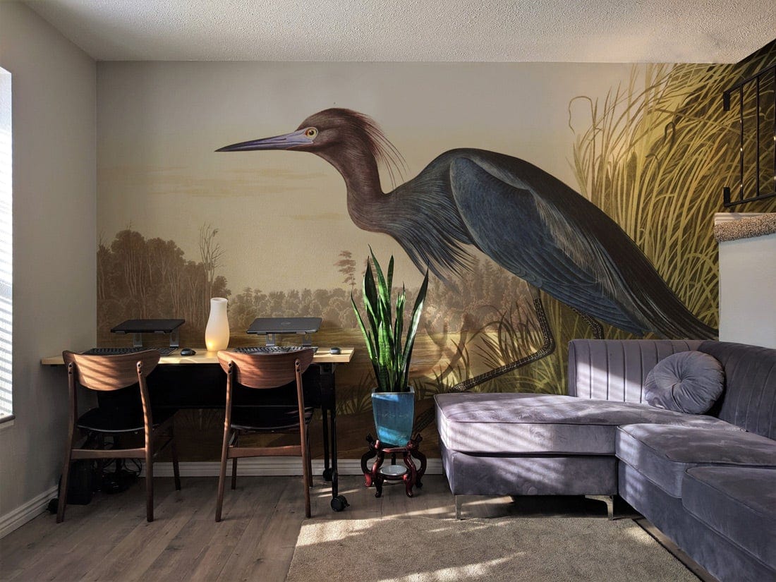 Elegant Blue Heron mural wallpaper, grey corner sofa, wooden chairs, modern room with natural lighting.