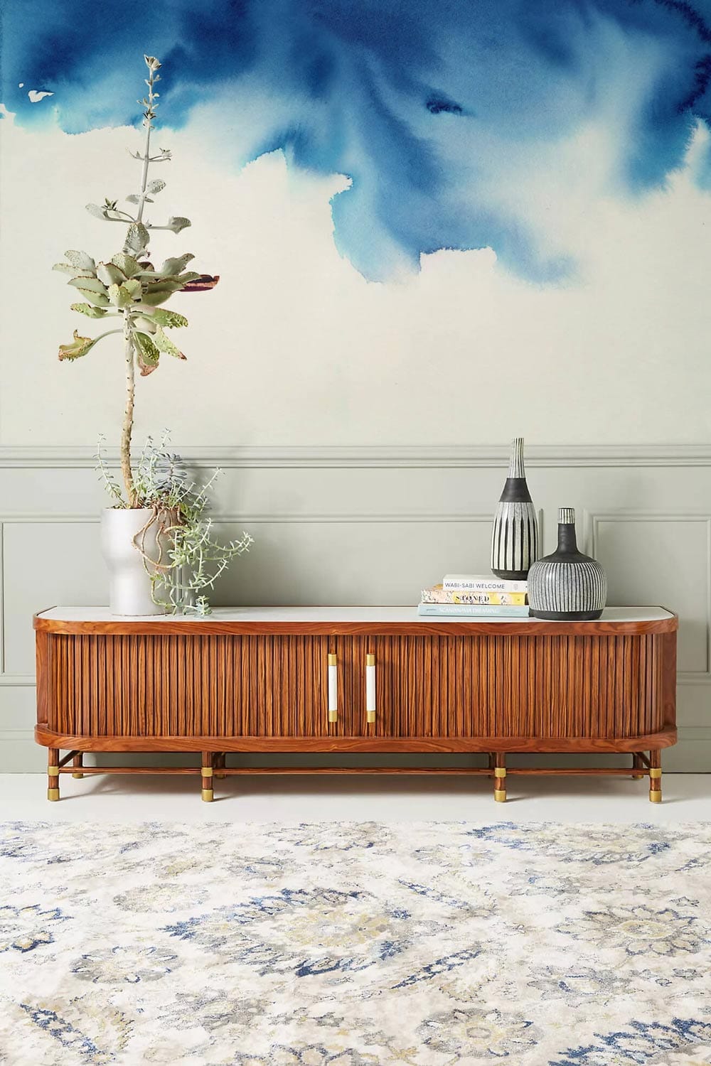 Elegant blue watercolour sky mural wallpaper with vintage wooden sideboard, patterned rug in modern interior.