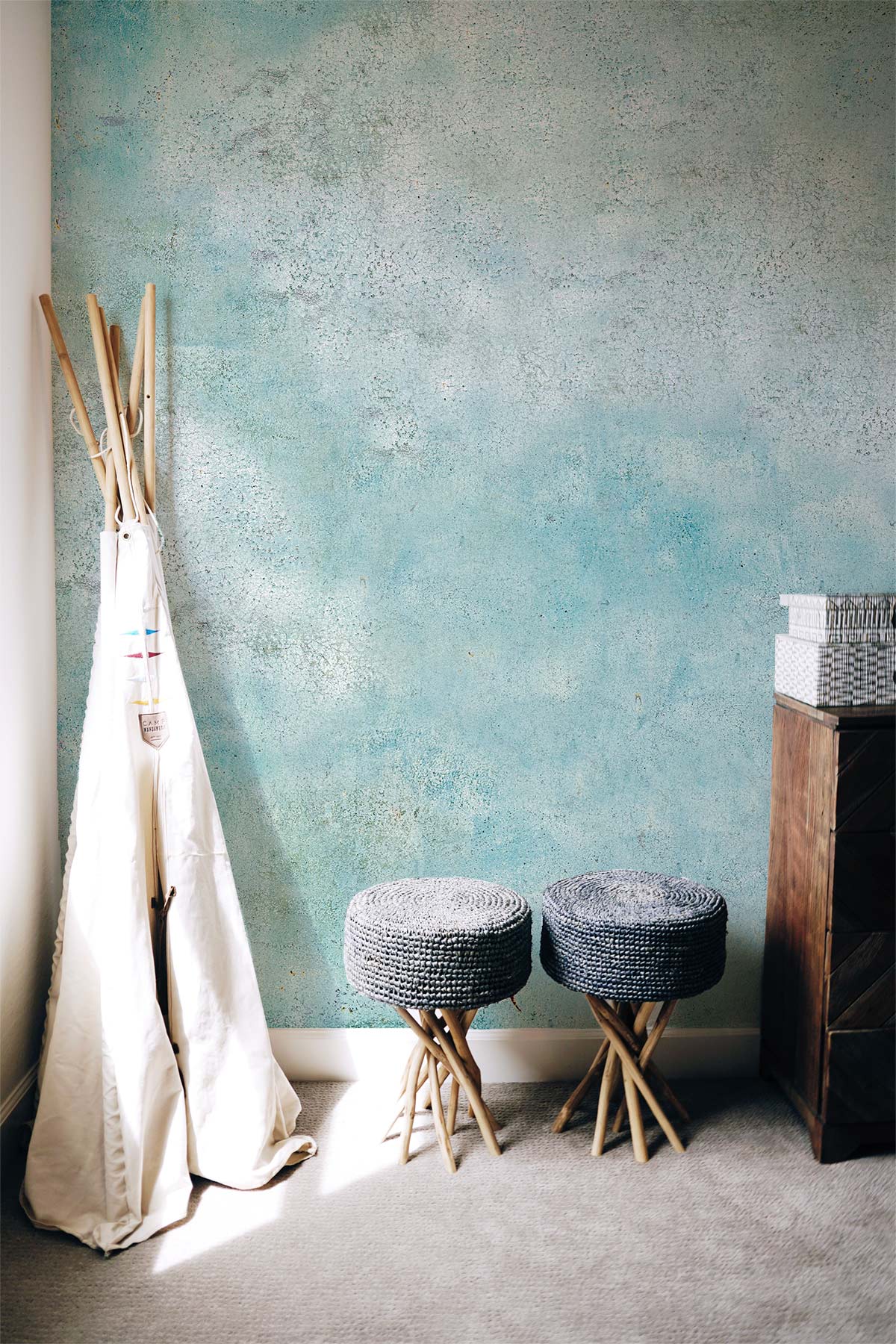 Vintage blue textured mural wallpaper with distressed aqua design, two woven stools, wooden cabinet, carpet flooring.
