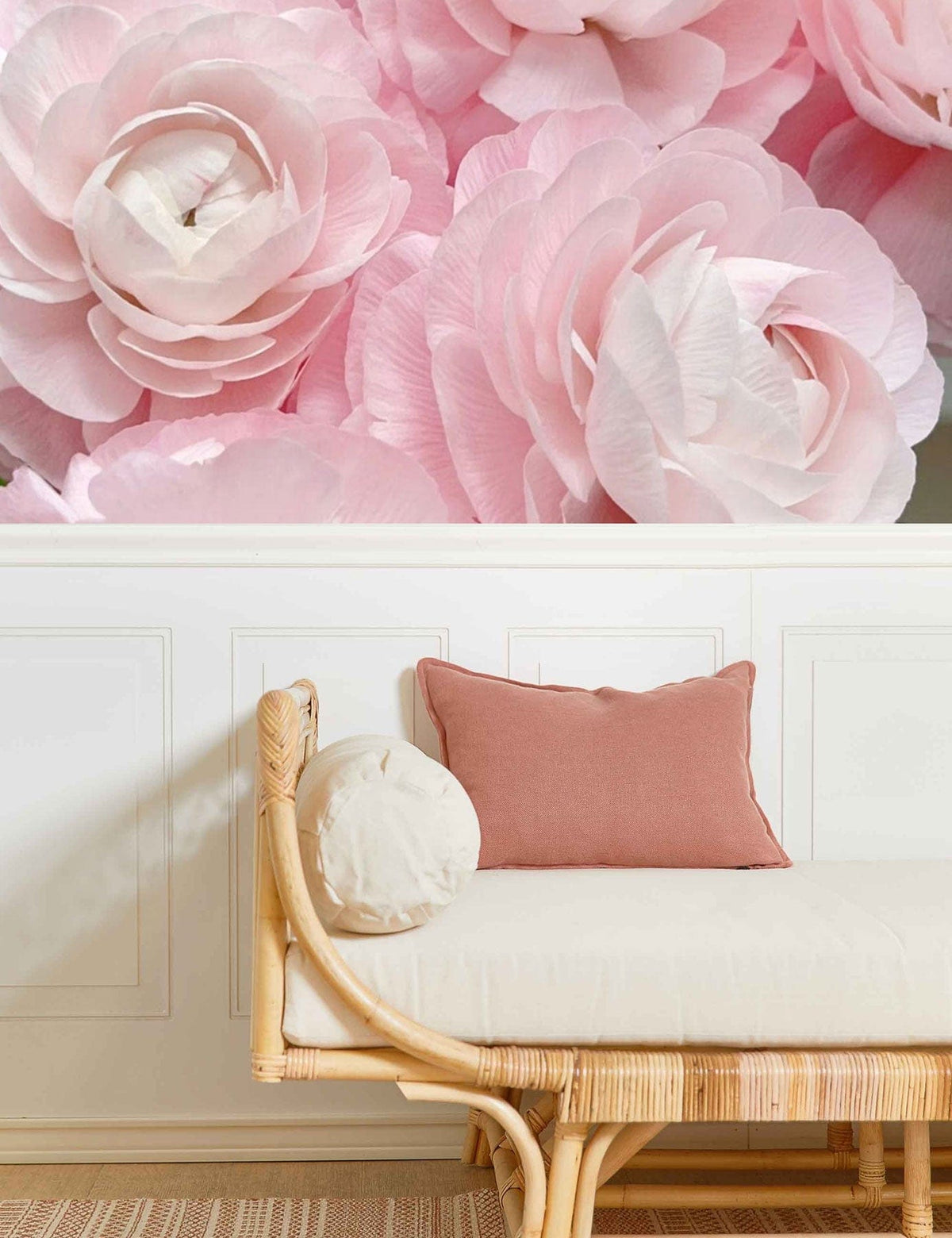 Blush pink ranunculus wallpaper, white wall panelling, wicker sofa, pink cushions, beige carpet flooring.