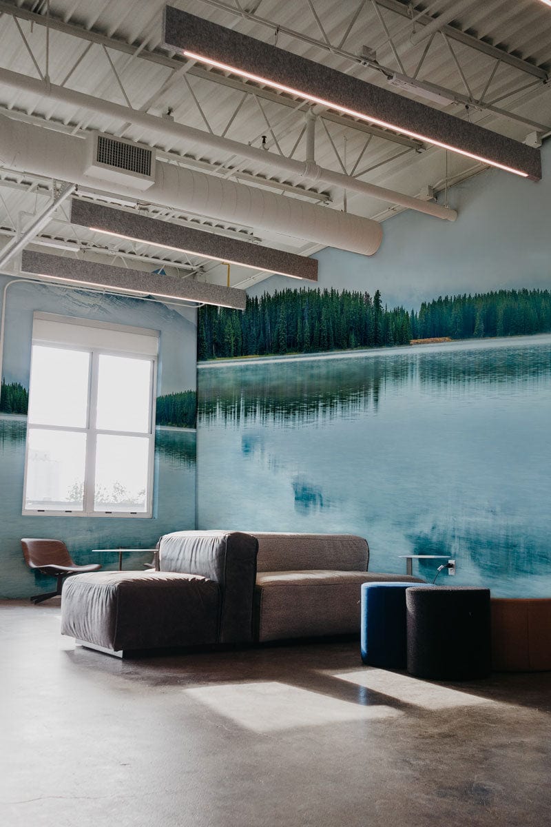 Tranquil blue mountain mural wallpaper, modern grey sofa, industrial ceiling, polished concrete flooring, office setting.