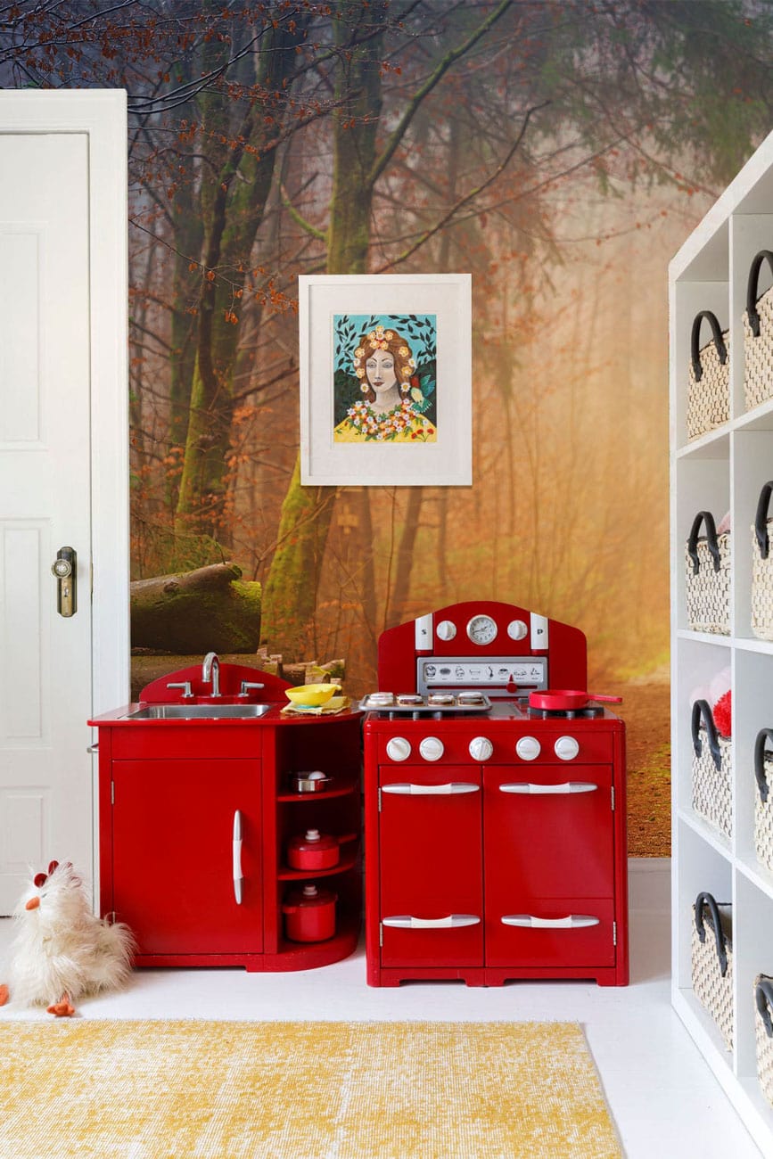 Red toy kitchen set against misty autumn woodland mural, white shelving with baskets, yellow rug.