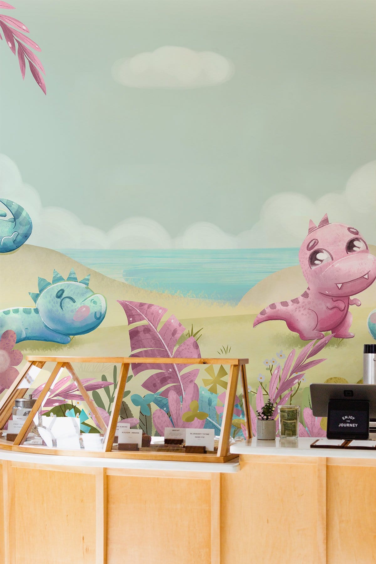 Nursery mural with playful pastel dinosaurs, light wooden counter, and coastal scenic background.