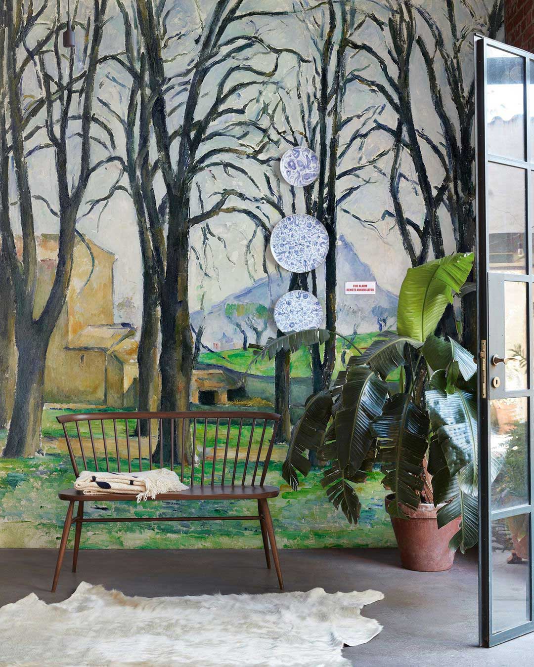 Vintage landscape mural wallpaper in rustic room with wooden bench, grey flooring, and large green plant.