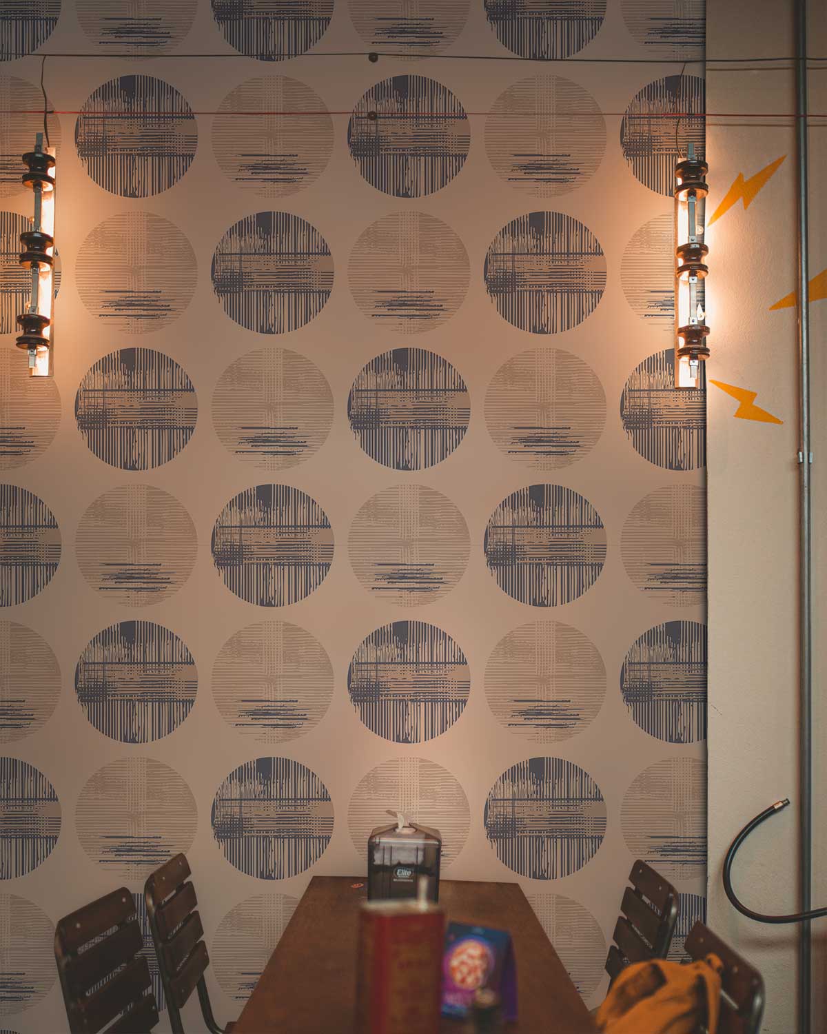 Modern geometric wallpaper with blue-beige circles, wooden chairs, industrial lights, and table in dining room.