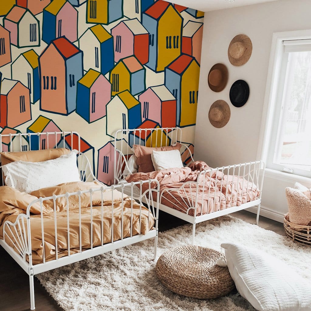 Colourful geometric houses wallpaper in cosy bedroom, white metal beds, beige rug, woven wall decor.