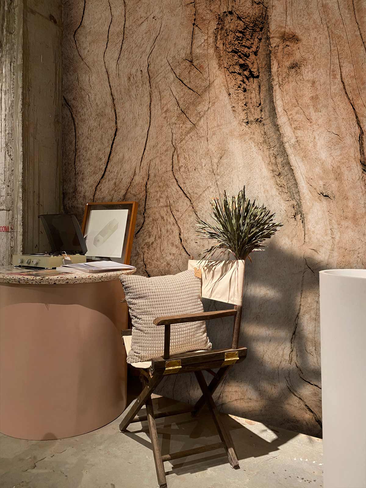 Rustic wood grain wallpaper, natural tones, round table, wooden folding chair, concrete flooring in cosy corner.