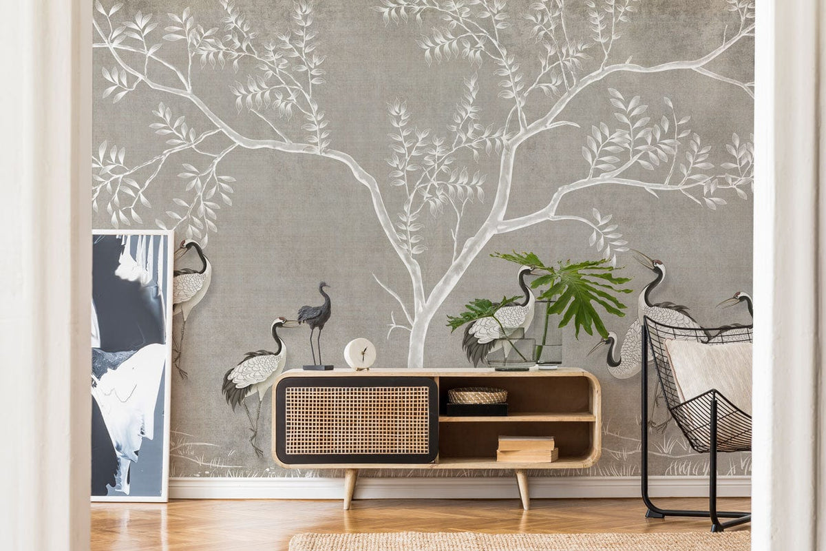 Elegant grey mural wallpaper with cranes and trees, wooden sideboard, wicker chair, art piece.