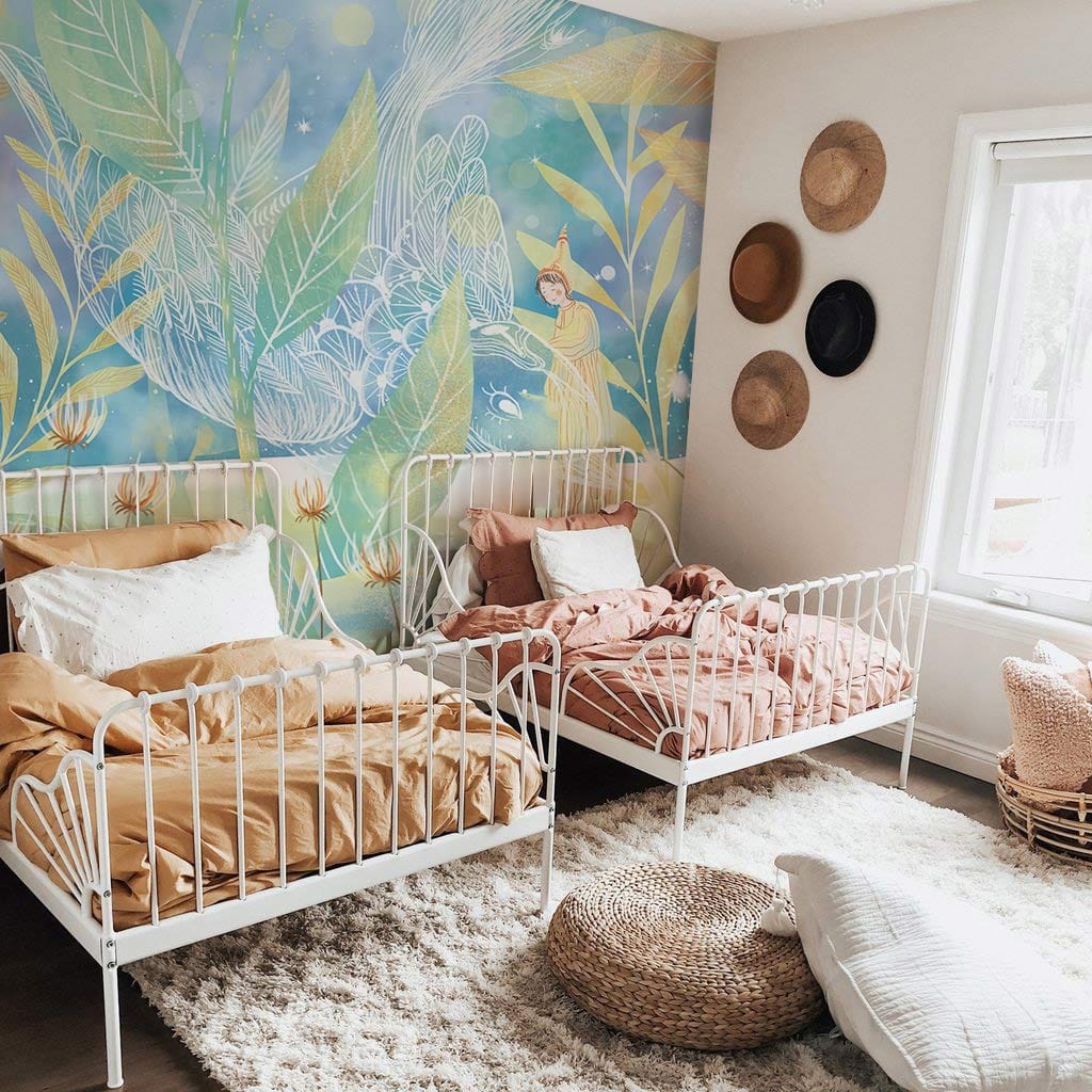 Children's bedroom with pastel forest mural, white beds, beige walls, shaggy rug, wicker decor items.