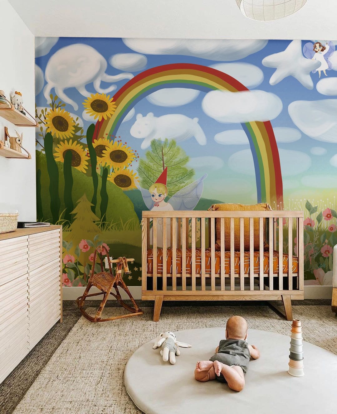 Whimsical nursery with fairyland mural, rainbow, sunflowers, wooden cot, rocking horse, neutral flooring.