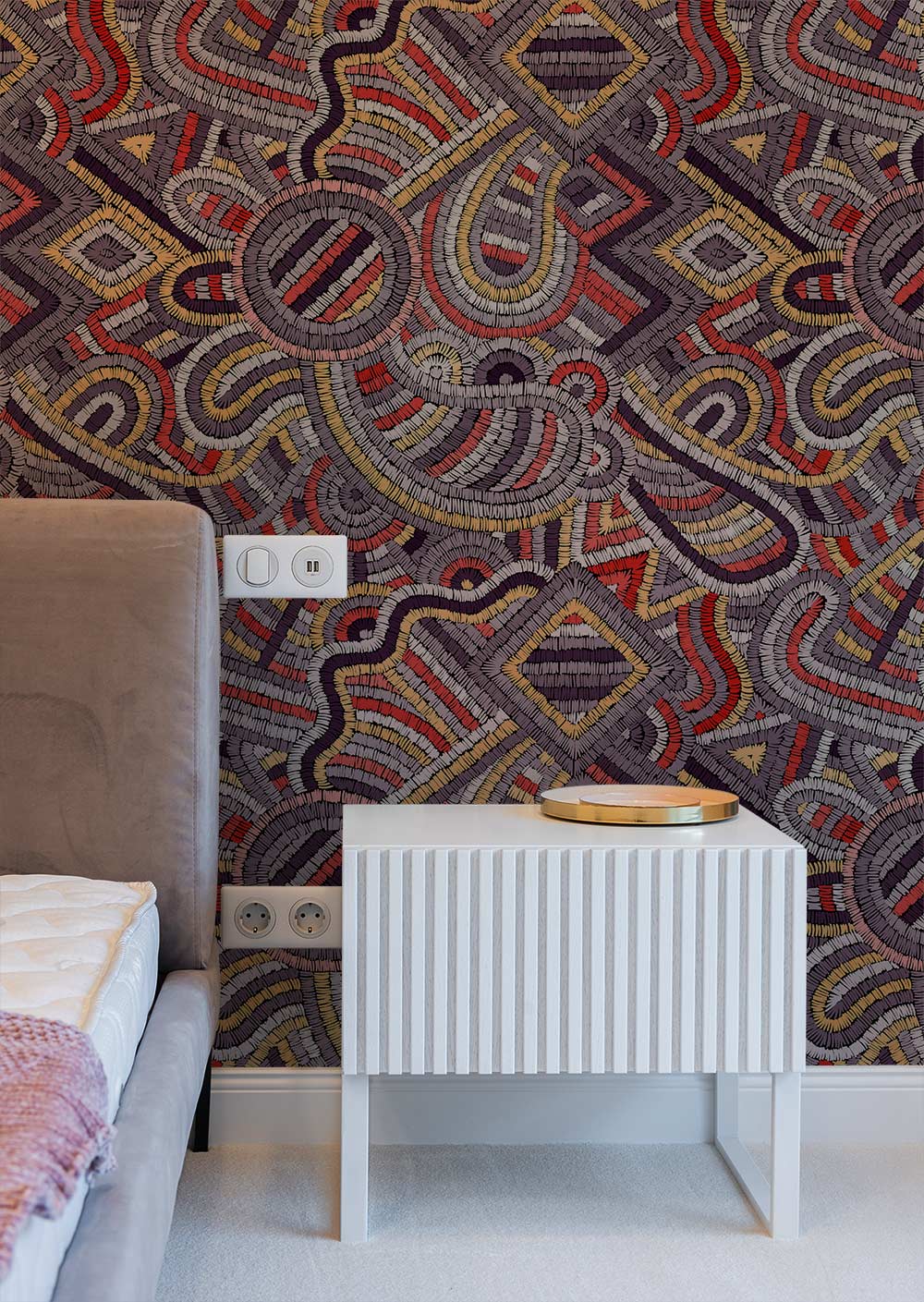 Multicoloured geometric wallpaper with tribal design alongside beige sofa and white side table in bedroom.