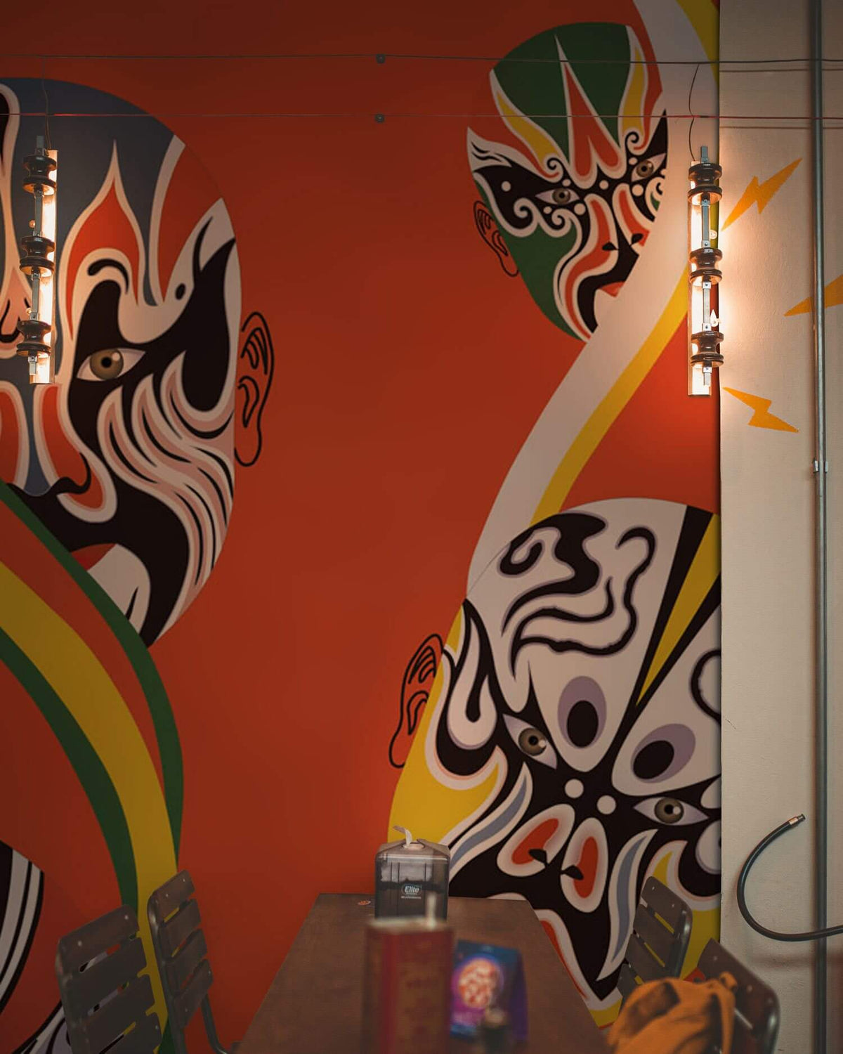 Vibrant Oriental Mask Pattern Wallpaper in dining room with industrial lights and wooden chairs. Bold design.