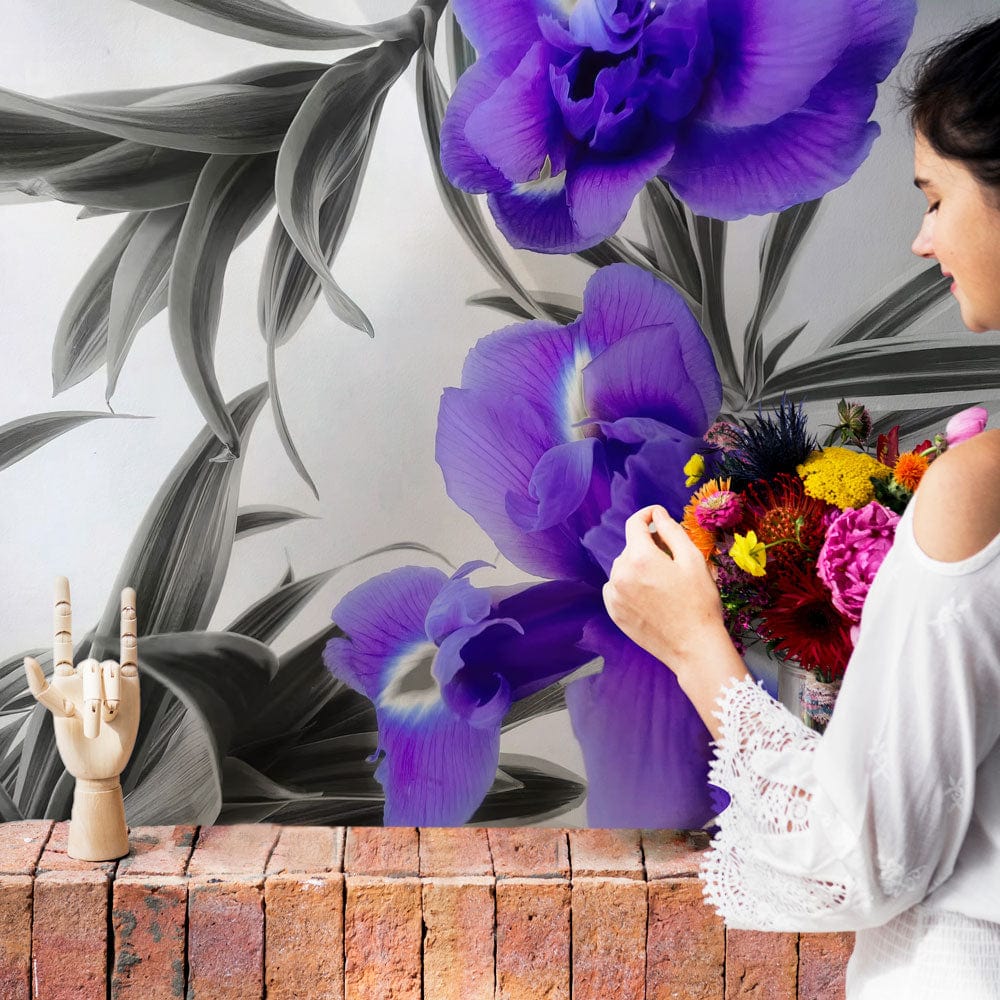 Vibrant purple floral mural wallpaper with elegant grey leaf accents behind woman holding flowers indoors.