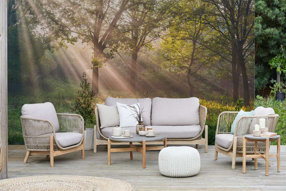 Outdoor seating with beige sofa and chairs, wooden deck flooring, forest landscape wallpaper backdrop.