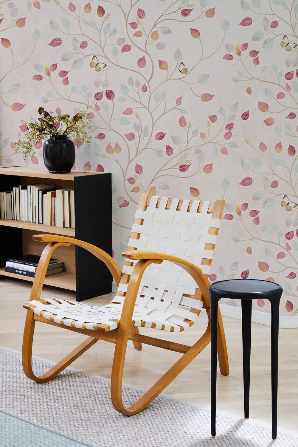 Vintage floral pastel mural wallpaper, armchair, bookshelf, light wood flooring, contemporary black side table.