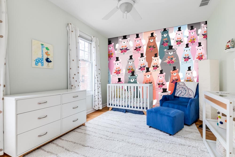 Charming Bears nursery wallpaper with crib, blue armchair, white furniture, light rug in child's bedroom.