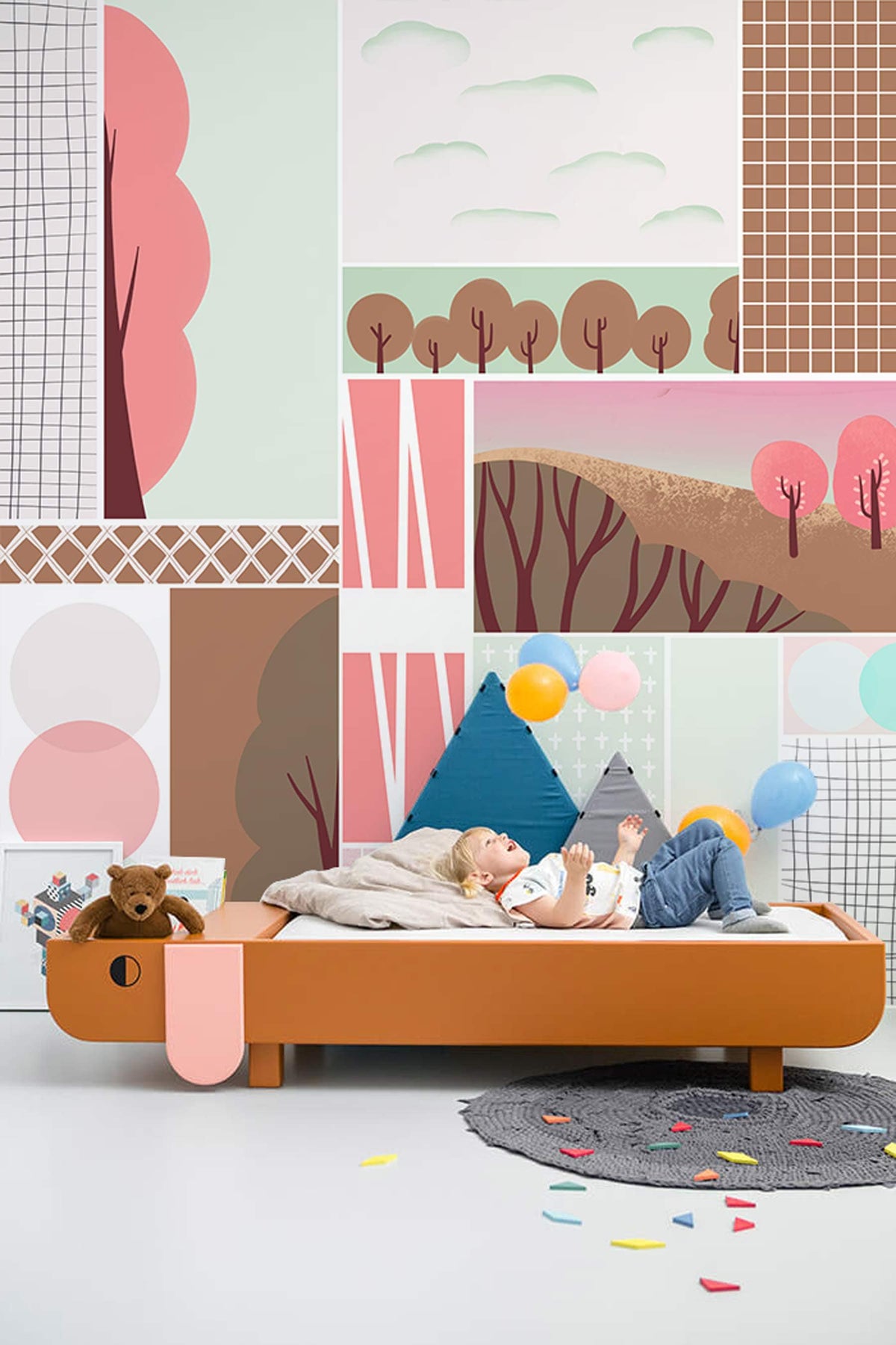 Pastel geometric landscape mural wallpaper in children's room with wooden bed, colourful balloons, grey rug.