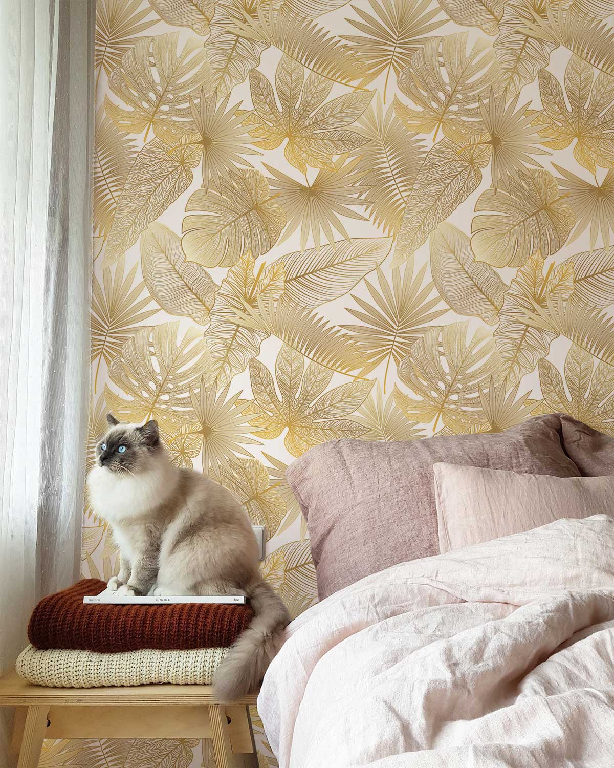 Elegant gold tropical leaf wallpaper, cosy bedroom, blush linens, wooden stool, cat, white curtains.