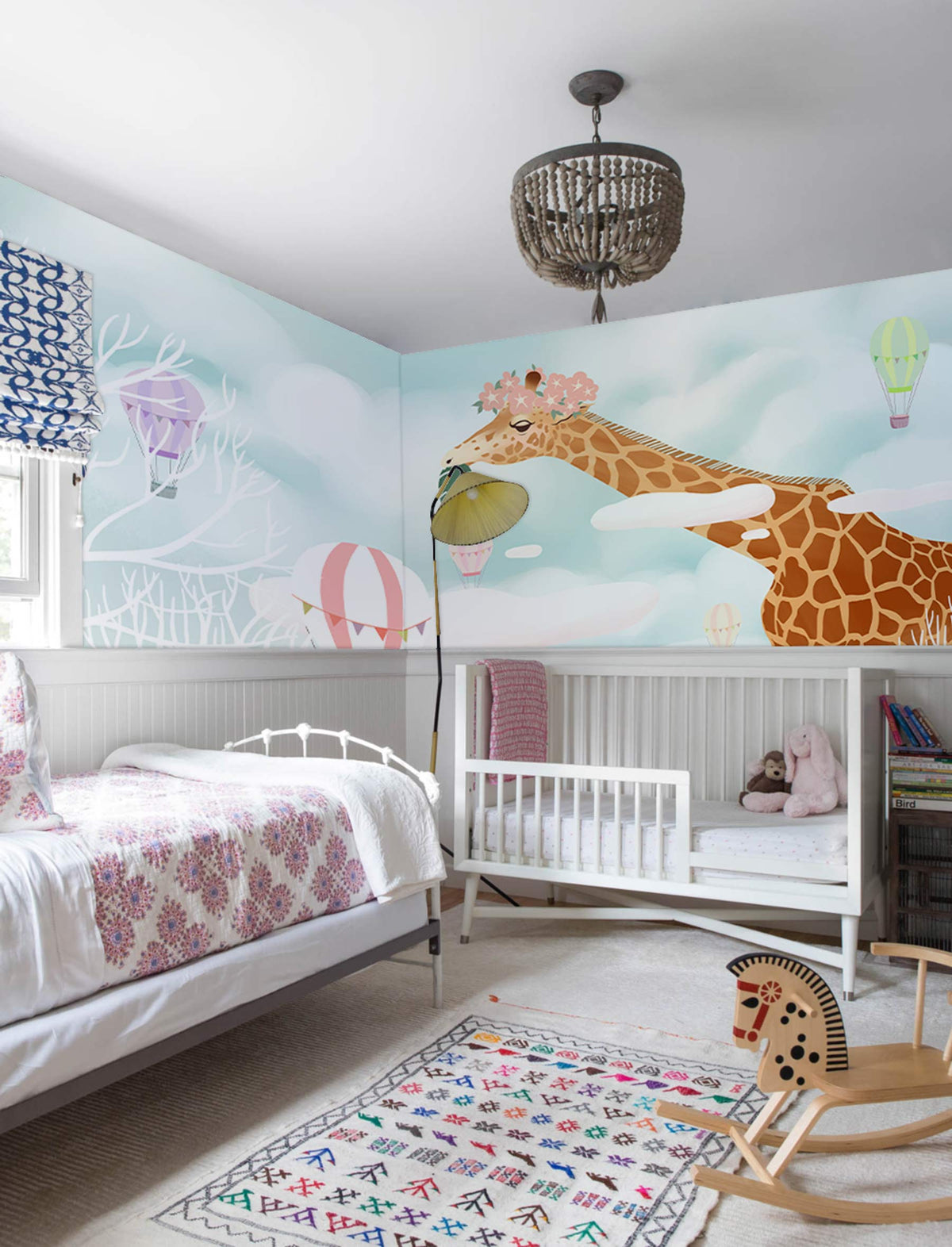 Whimsical Giraffe mural in pastel nursery with crib, white walls, patterned rug, and ceiling chandelier.