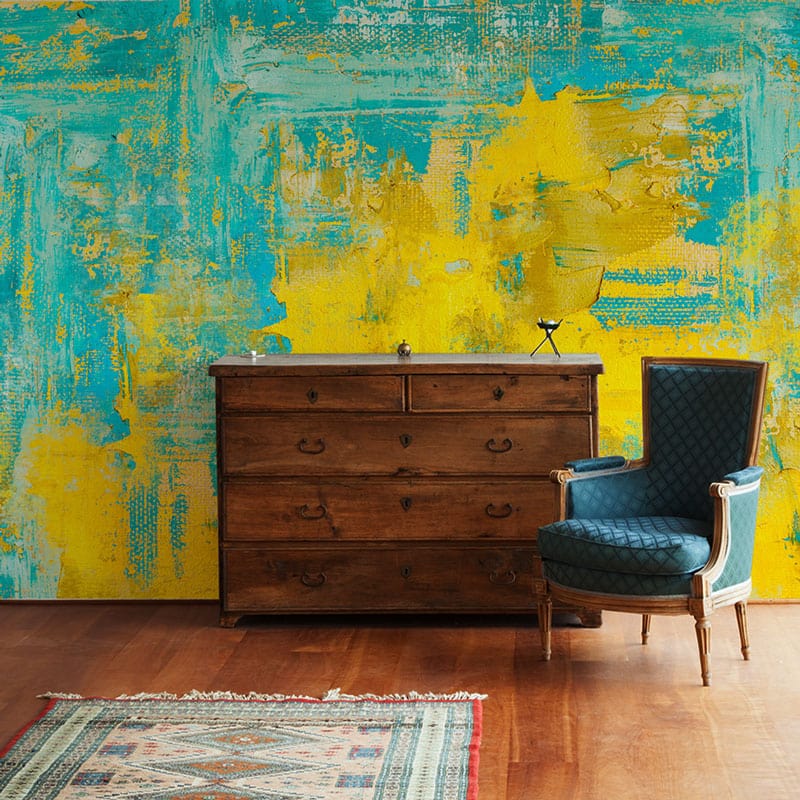 Teal yellow abstract mural wallpaper, wooden chest, blue armchair, wooden flooring, vibrant accent wall design.
