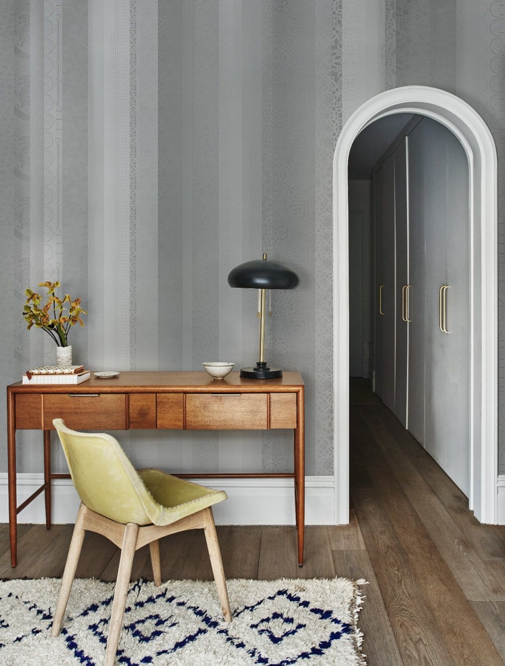 Mid-century desk and chair in grey room with arched doorway, patterned rug, wooden floor.