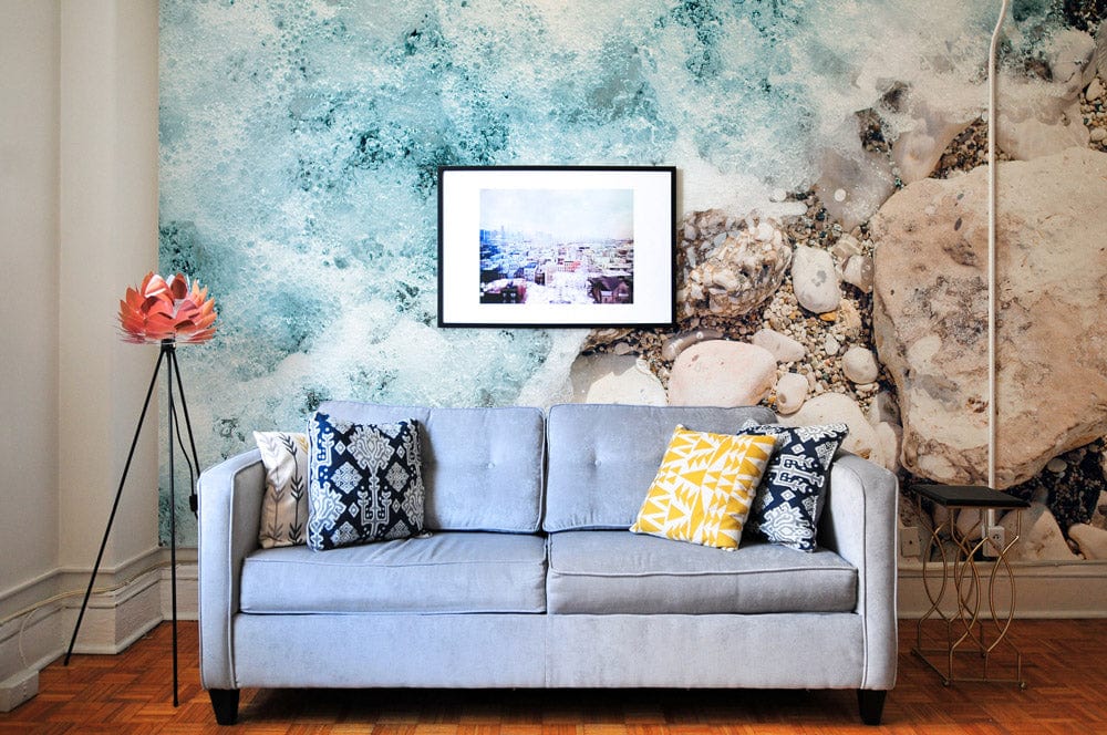 Coastal Serenity mural wallpaper with ocean foam, pebbles, blue-grey sofa, parquet flooring, and framed artwork.