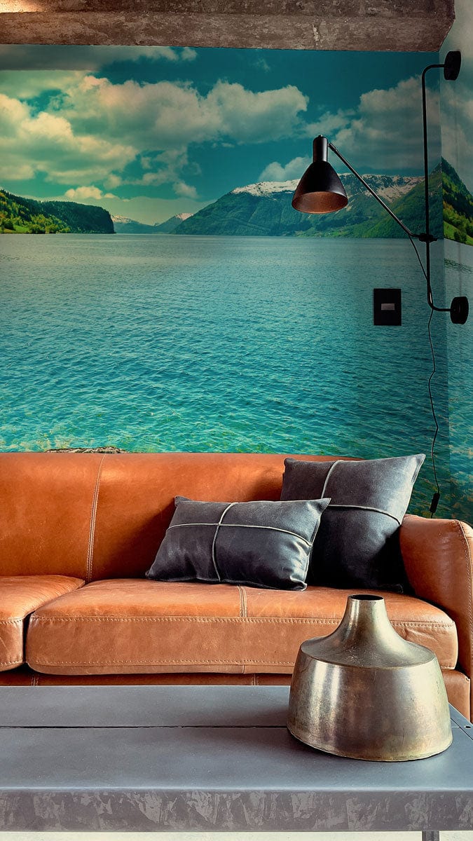 Tranquil blue lake mural wallpaper in living room with brown leather sofa, grey cushions, and lamp.