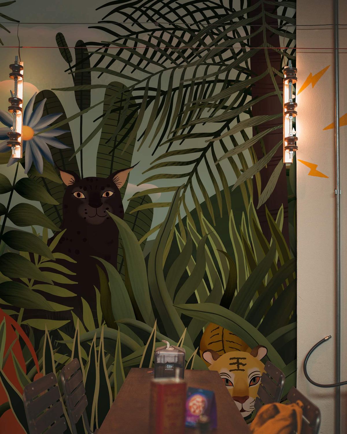 Colourful jungle animals mural wallpaper with tropical greenery; industrial lighting, dark dining table, wooden chair.