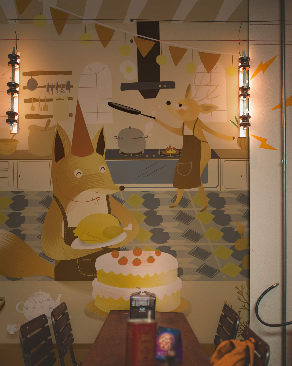 Whimsical woodland animal bakery mural wallpaper in soft yellow tones with industrial pendant lights.