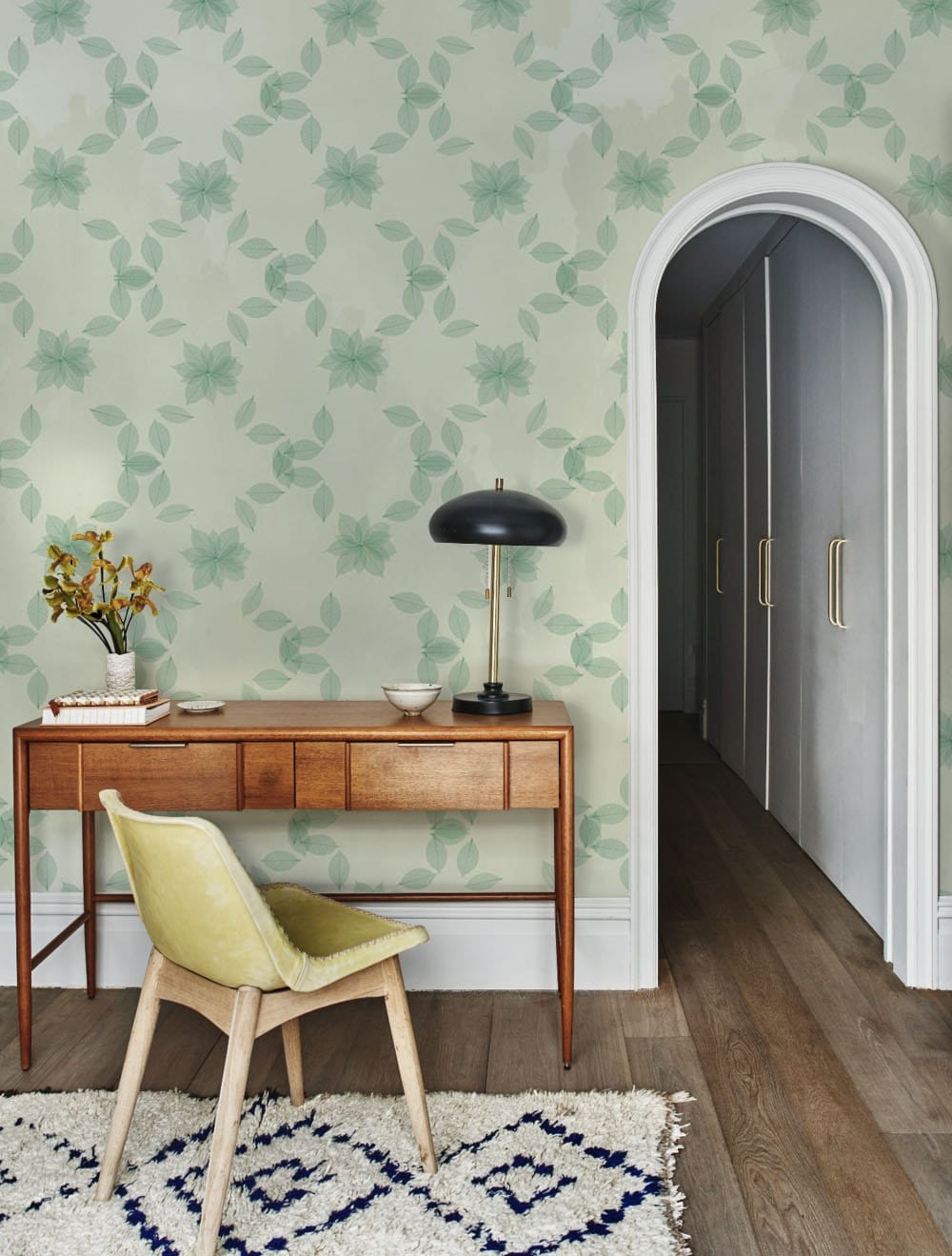 Elegant sage green floral wallpaper in hallway with mid-century desk, velvet chair, wooden flooring.