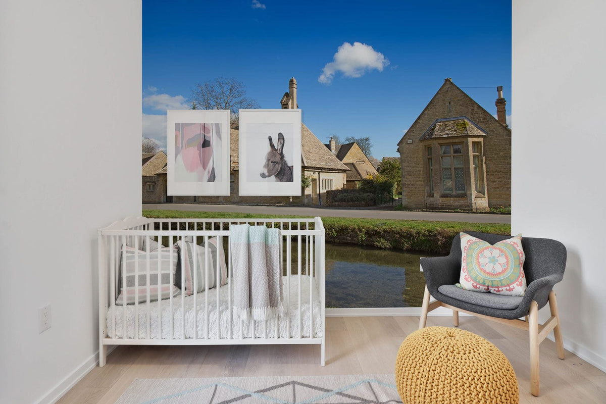 Charming Cotswold Village Scene Mural Wallpaper, nursery room, white cot, grey chair, woven pouf, light wood flooring.